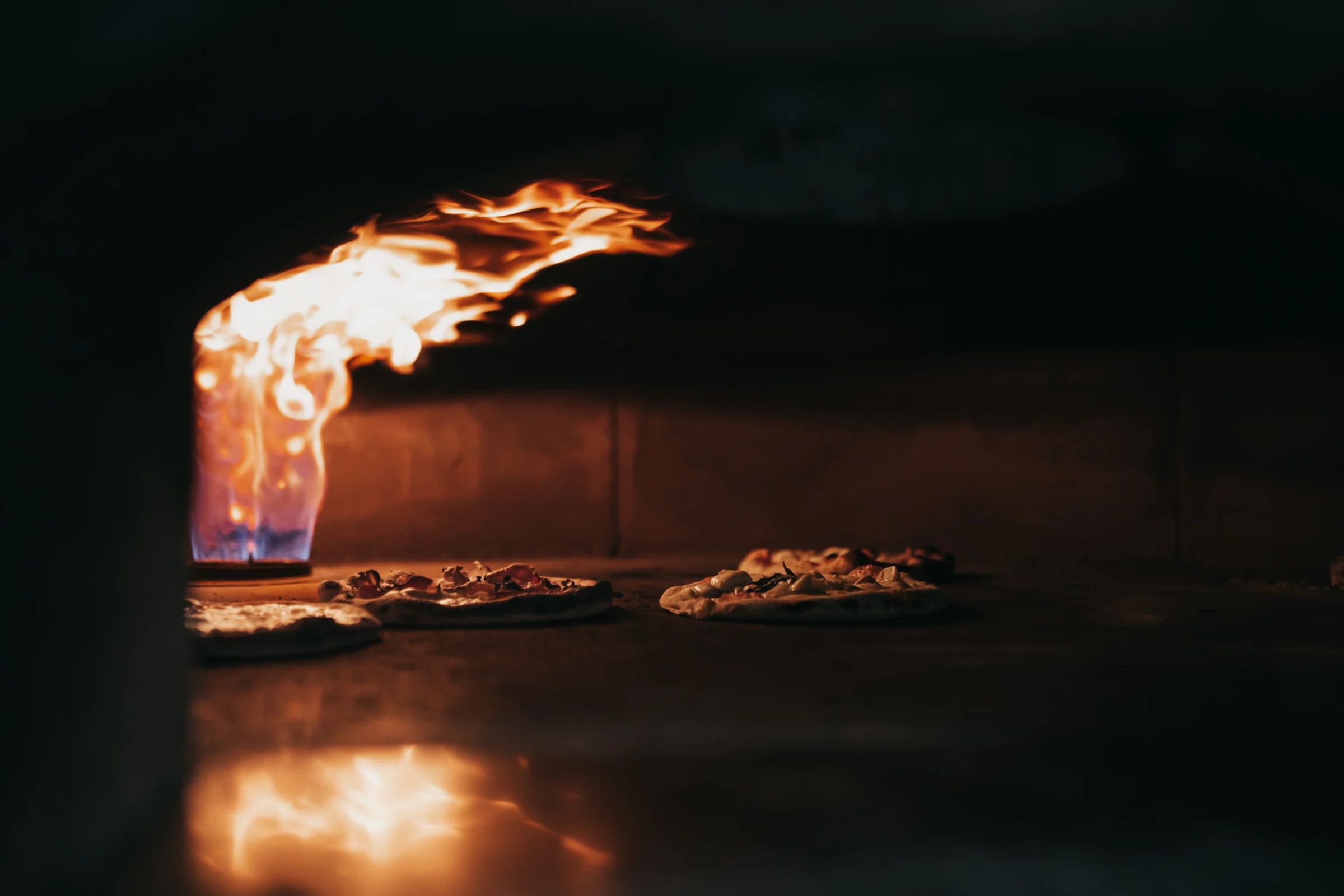 Tres pizzas sobre la piedra de un horno, con la llama curvándose por encima.